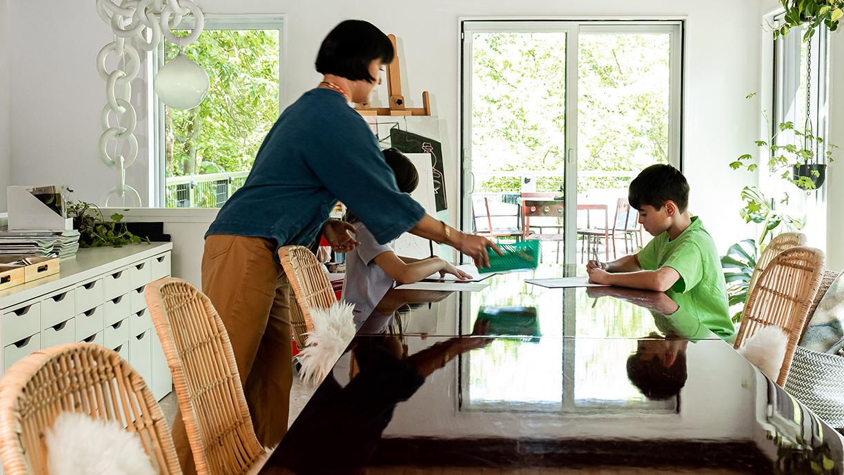 Inside a room with white walls and lots of windows, a woman sets a basket on a table where two children draw.