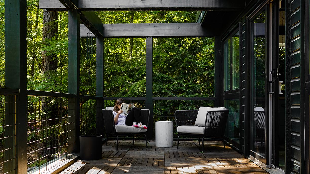 A little girl reads a book in a chair on a deck outside a home with a black exterior. 