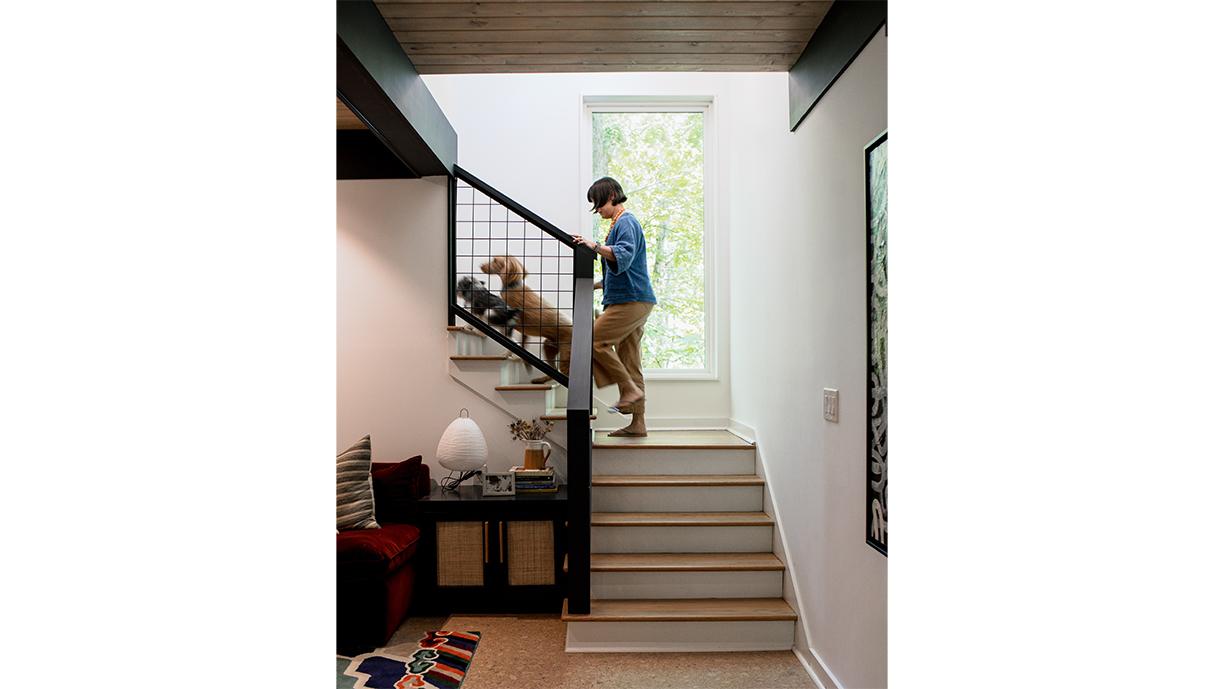 A women climbs a set of stairs with her two dogs passing in front of a large window set in the wall next to the stair landing.