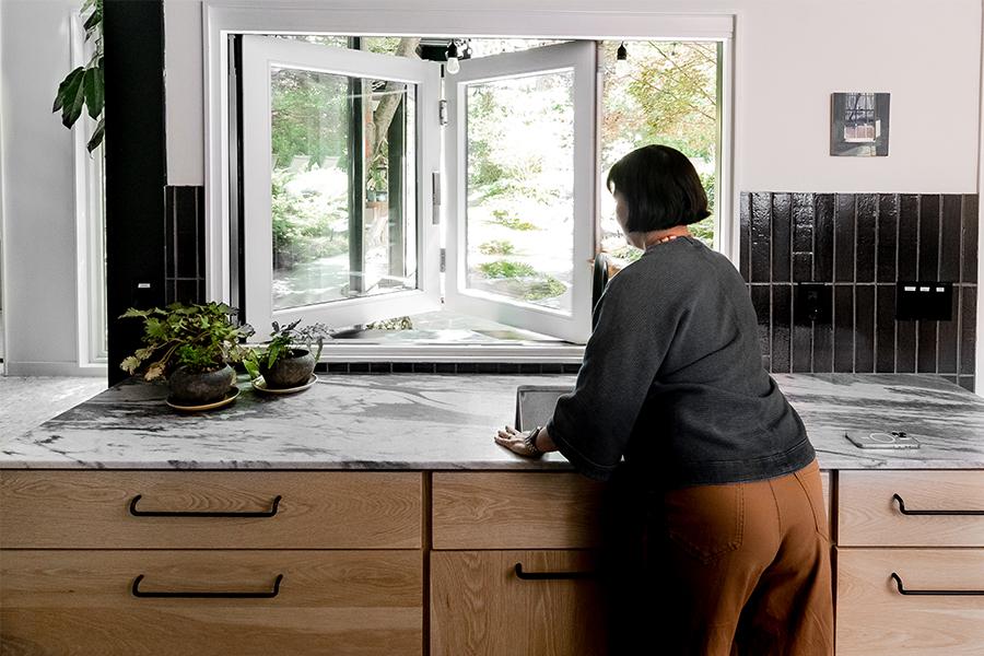 A woman reaches over a marble countertop and kitchen sink to open a folding pass-through window.