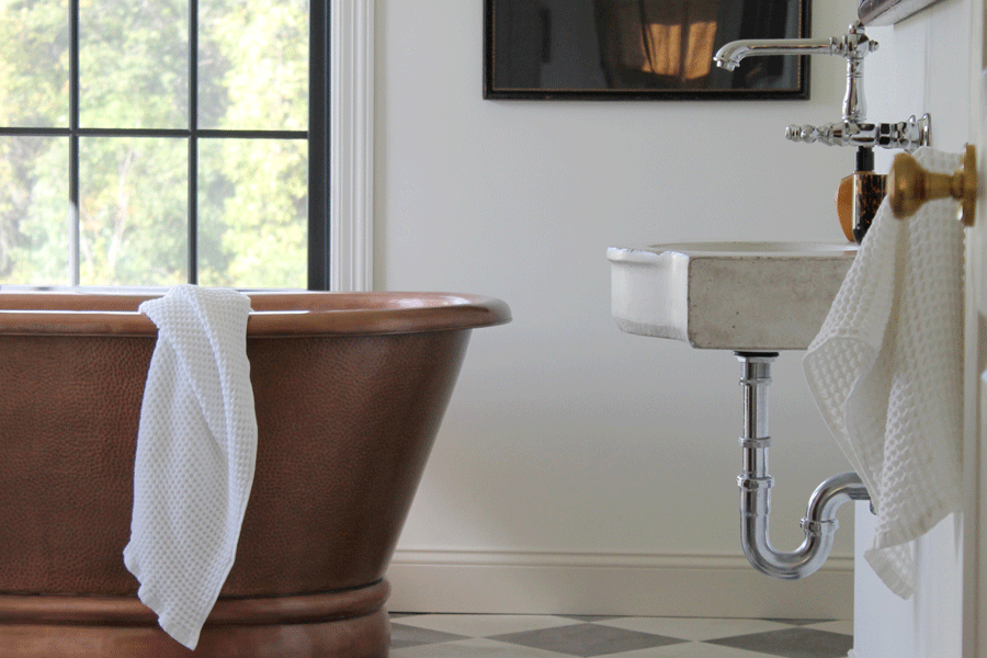 Two double-hung windows bring natural light into a bathroom with a copper tub and black-and-white checkerboard floor.