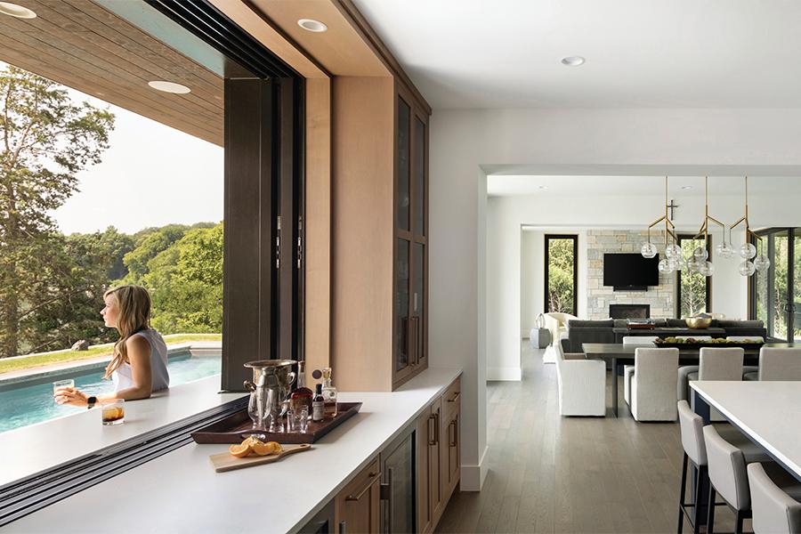 An open kitchen pass-through window sits flush with the countertop, which creates an outdoor bar where a woman sips her cocktail and looks out over the swimming pool. 