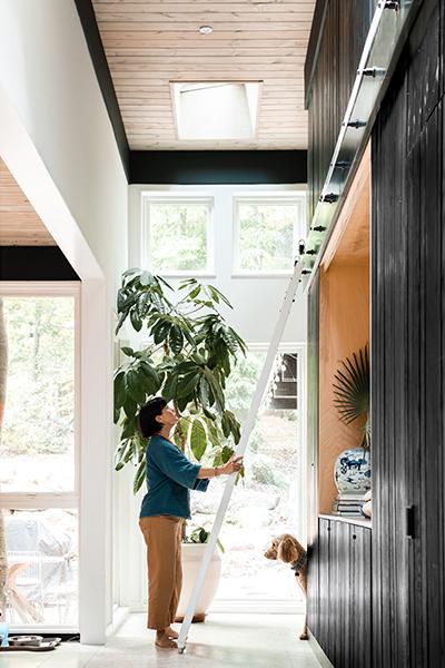 A women stands at the bottom of a ladder in a hallway lined with black wood paneling while light shines through windows at the end of the hall and a dog peeks around the corner.