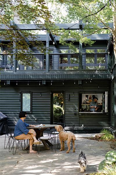 An exterior photo of a home showing a woman sitting at a table on a patio, two dogs milling about, and inside an open window, a man mixing a cocktail. 