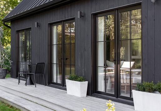 An exterior shot of a black barn-inspired addition featuring three sets of black E-Series Hinged Patio Doors and a deck with seating and potted plants.