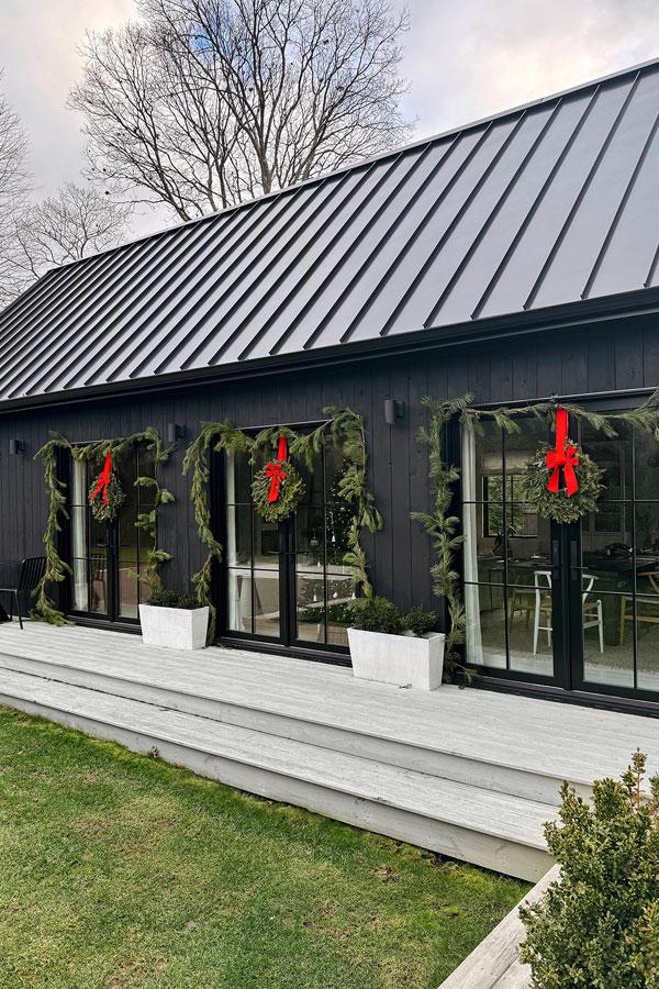 Two exterior shots of a black barn-inspired home addition with four sets of black E-Series Hinged Patio Doors that have been decorated for Christmas with evergreen boughs, wreaths, and red bows.