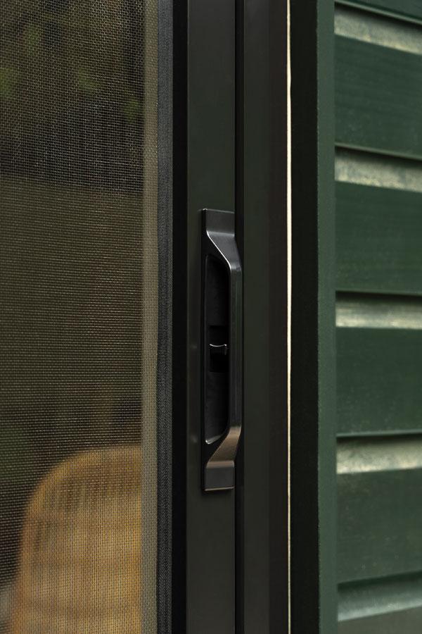 An exterior shot of a woman opening a black 200 Series Narroline® Gliding Patio Door and retractable insect screen to her deck where her dog awaits by a potted plant. 