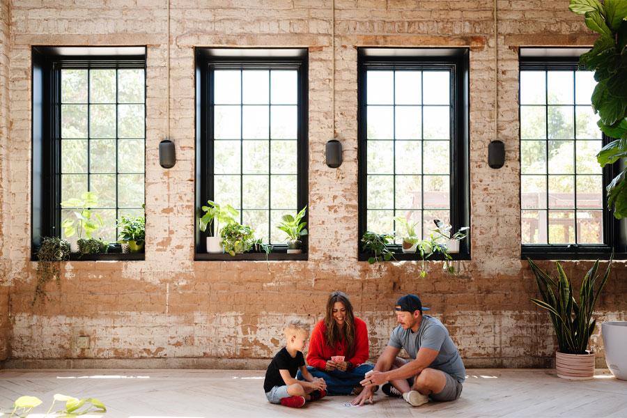 A family sits on the ground underneath a bank of four black windows and plays a game of cards.