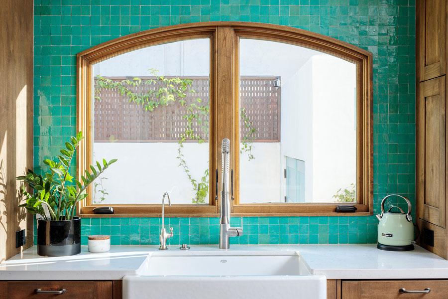 Two arch-top casement windows above a kitchen sink with a turquoise zellige tile backsplash and alderwood cabinetry.