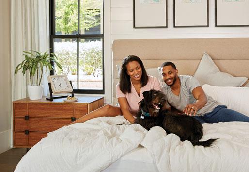 Couple pet their dog while relaxing in bed with double-hung windows behind them