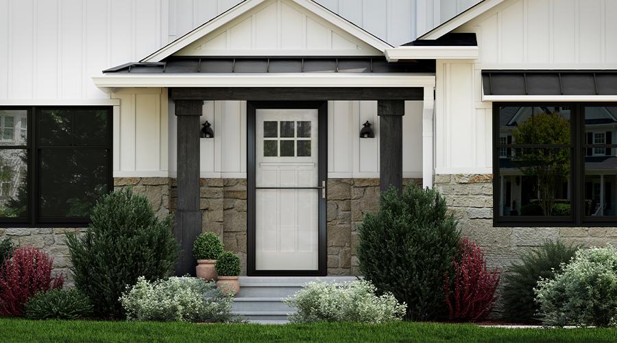 An exterior view of a home’s entryway showing its white siding, black windows, landscaping around the front door, and a black storm door over a white front door.