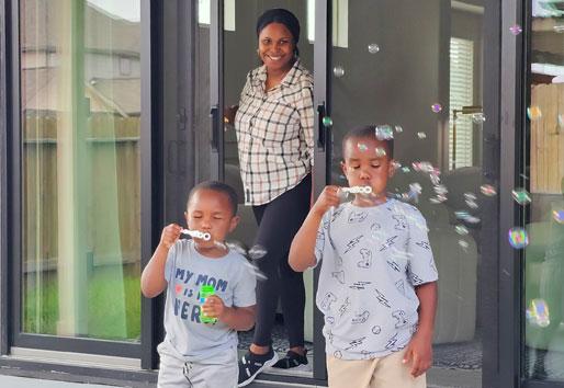 A woman is standing in the doorway of her patio door closing a retractable screen. Her two children are standing in front of her blowing bubbles on the patio. 