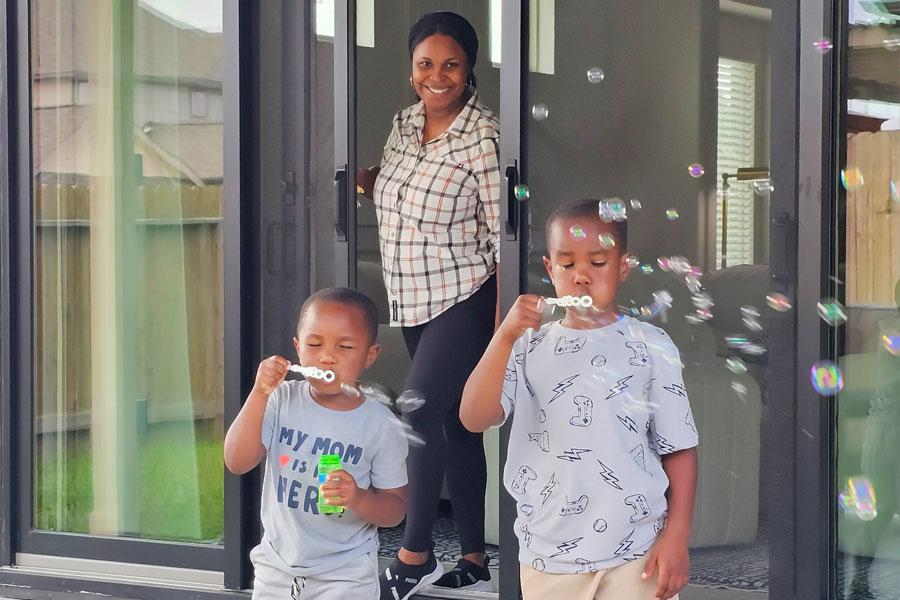 A woman is standing in the doorway of her patio door closing a retractable screen. Her two children are standing in front of her blowing bubbles on the patio. 